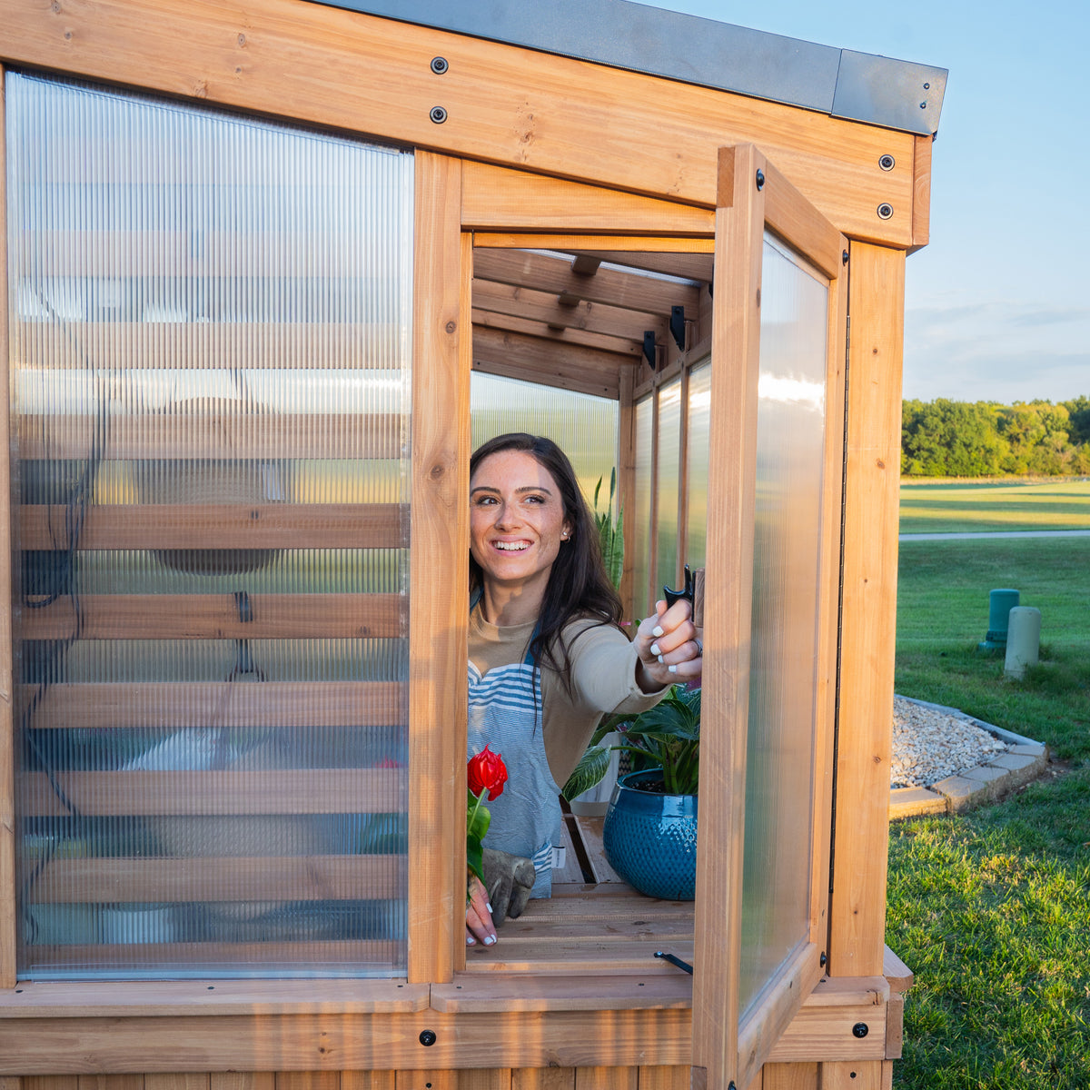 Woman opening the wide entry door of the Willow 9x6 greenhouse kit with potted plants on the interior shelf.