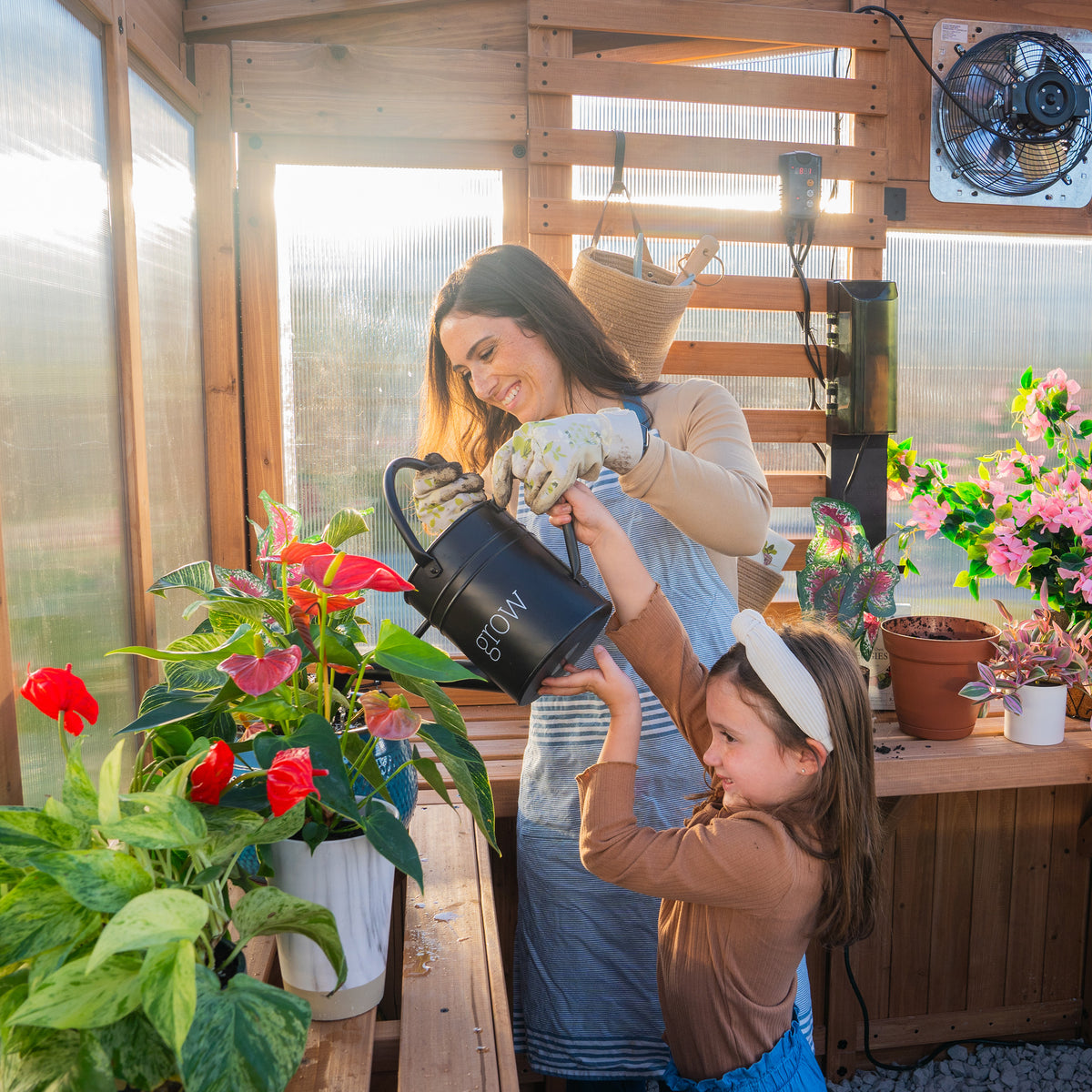 A woman and child water potted plants together on the staging shelves inside the Willow cedar greenhouse.
